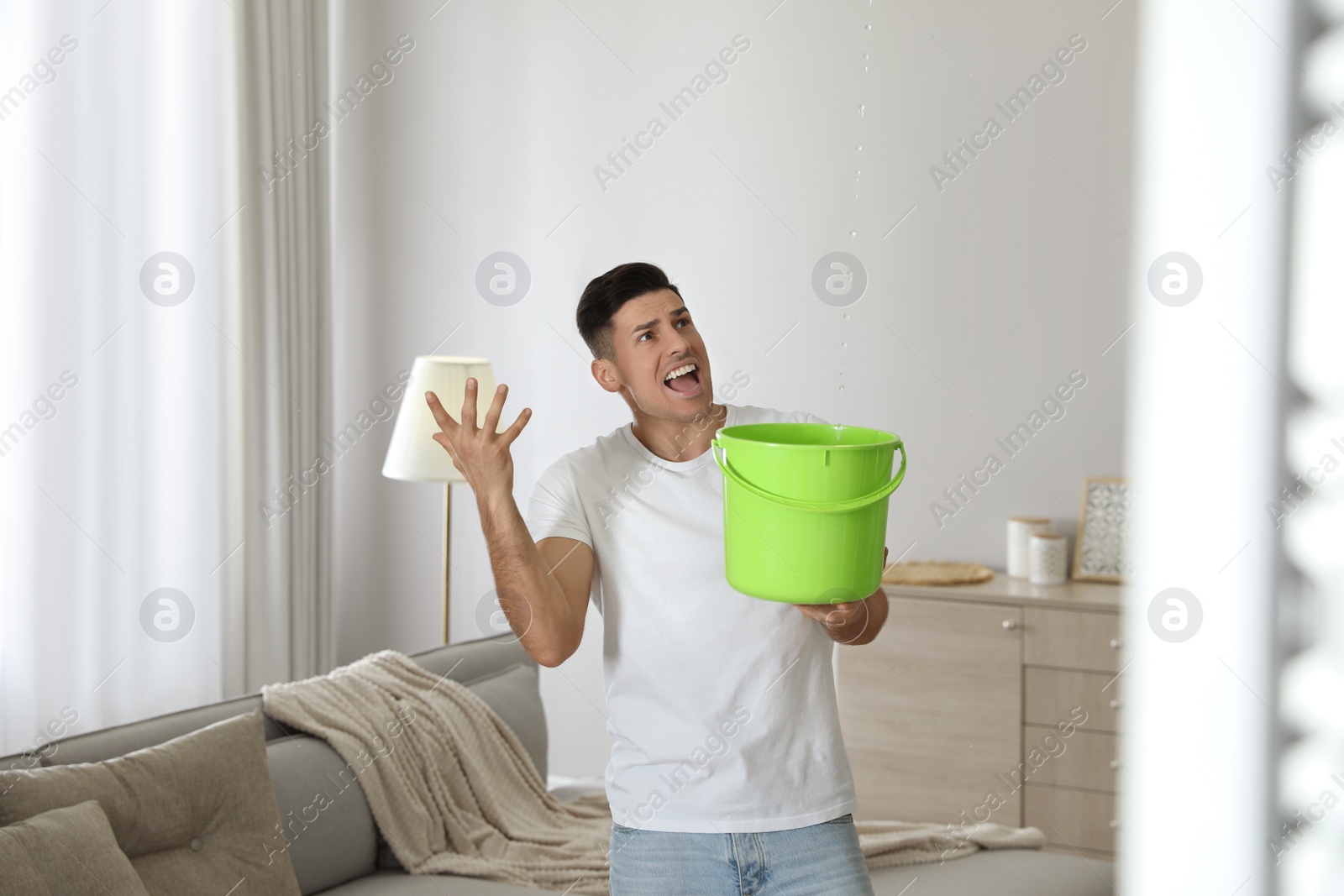 Photo of Emotional man collecting water leaking from ceiling in living room. Damaged roof