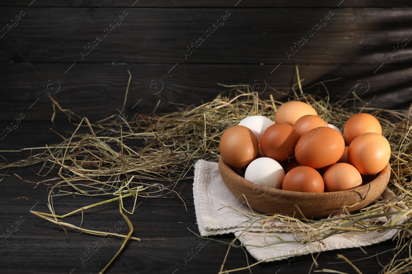 Photo of Fresh chicken eggs in bowl and dried hay on black wooden table. Space for text