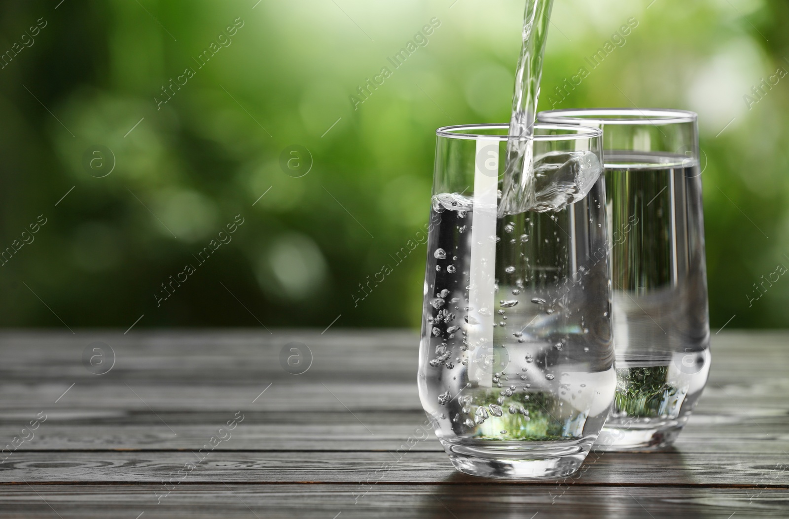 Photo of Pouring water into glass on wooden table outdoors, space for text