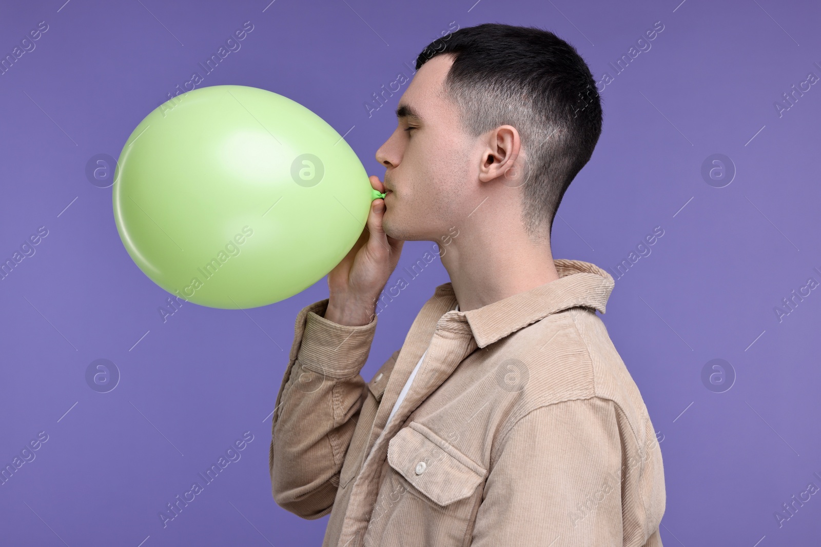 Photo of Young man inflating light green balloon on purple background