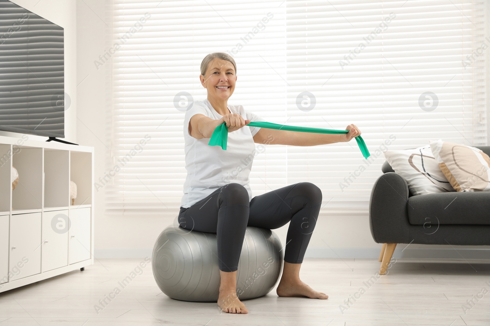 Photo of Senior woman doing exercise with elastic resistance band on fitness ball at home