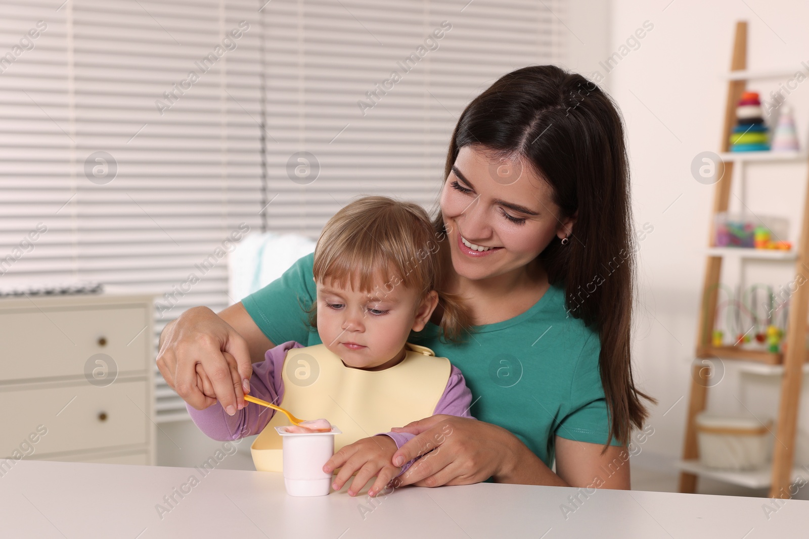 Photo of Mother feeding her cute little child with yogurt at white table in room