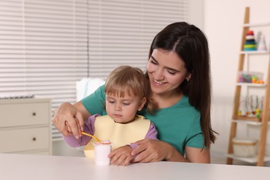 Mother feeding her cute little child with yogurt at white table in room