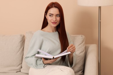 Photo of Beautiful woman with red dyed hair and book sitting on sofa indoors