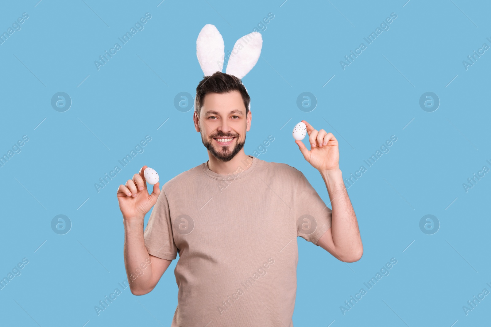 Photo of Happy man in bunny ears headband holding painted Easter eggs on turquoise background