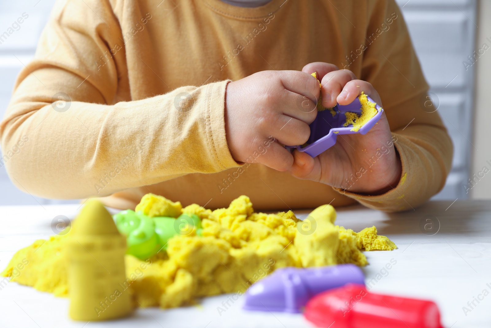 Photo of Little child playing with yellow kinetic sand at white table, closeup