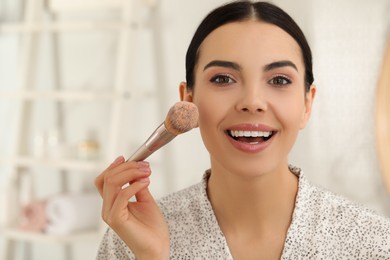 Beautiful young woman applying face powder with brush in bathroom at home