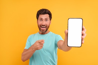 Handsome man showing smartphone in hand and pointing at it on yellow background