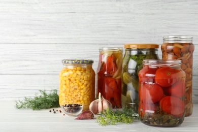 Glass jars with pickled vegetables on white table against wooden background