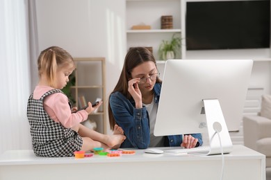 Photo of Woman working remotely at home. Tired mother using computer while her daughter playing with phone. Child sitting on desk