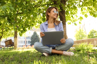 Happy young woman with laptop sitting on green grass in park