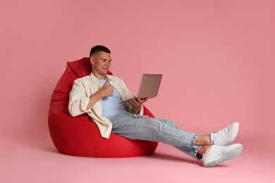 Photo of Handsome man with laptop showing thumb up on red bean bag chair against pink background