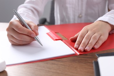 Woman writing on sheet of paper in red folder at wooden table in office, closeup