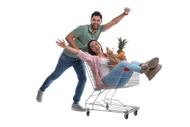 Photo of Young man giving his girlfriend ride in shopping cart with groceries on white background