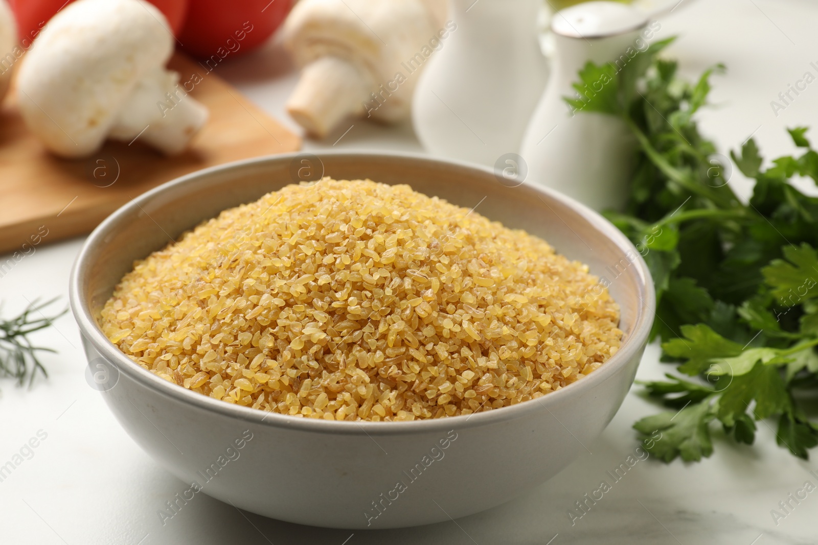 Photo of Raw bulgur in bowl and spices on white table, closeup