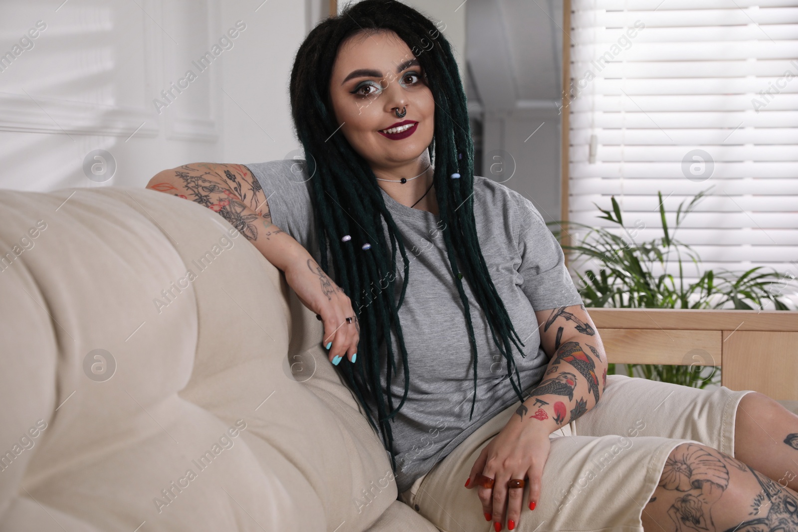 Photo of Beautiful young woman with tattoos on body, nose piercing and dreadlocks at home