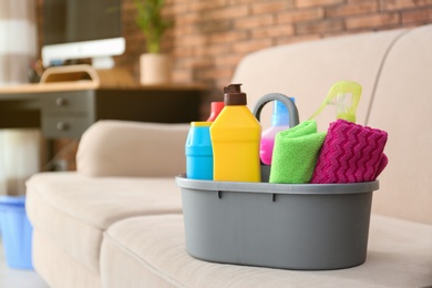 Photo of Basin with different detergents on sofa indoors, space for text. Cleaning service
