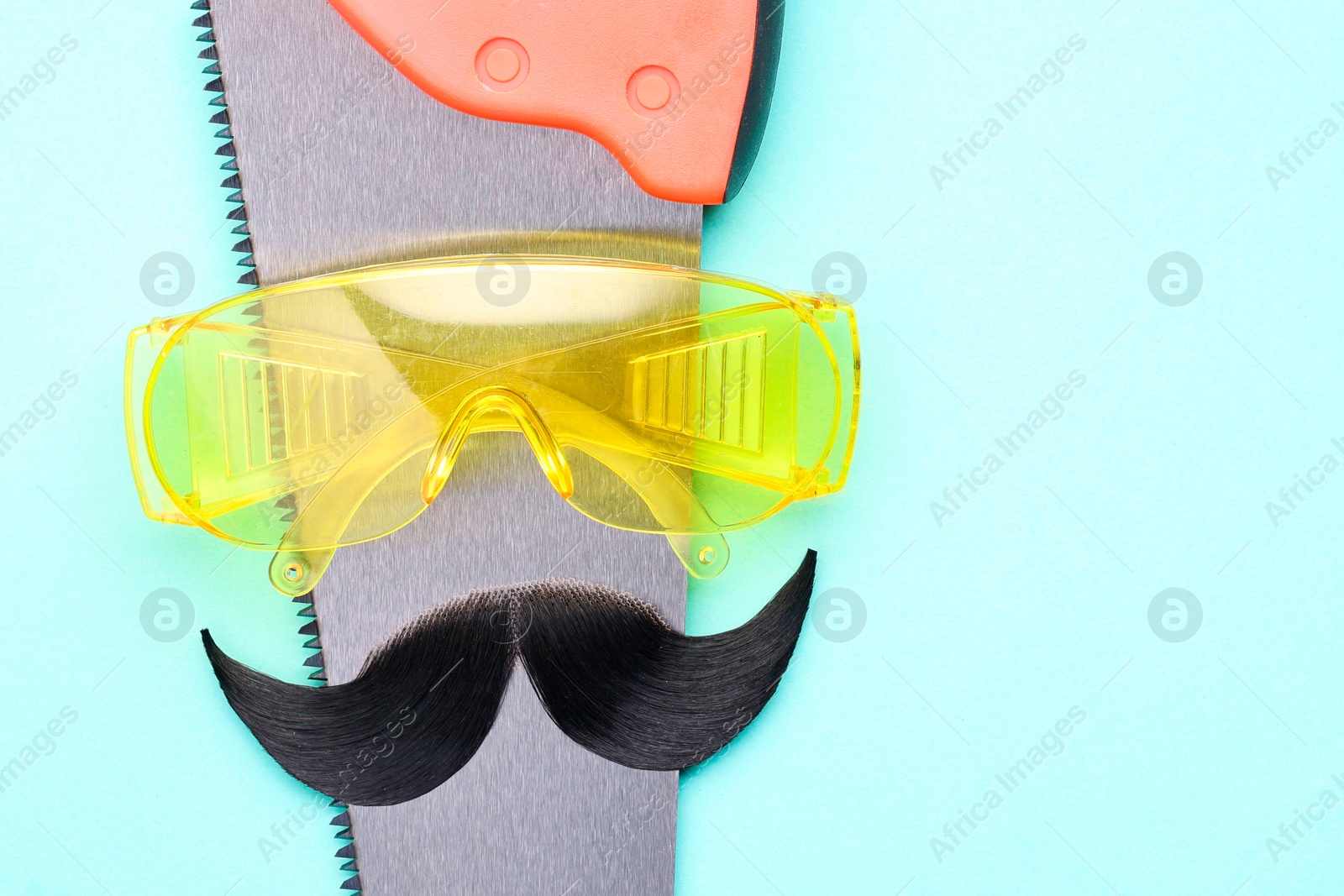 Photo of Man's face made of artificial mustache, safety glasses and hand saw on light blue background, top view. Space for text