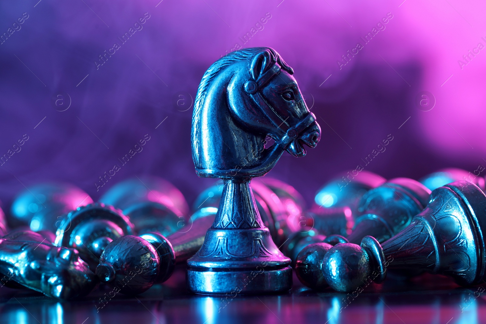 Photo of Metal knight among fallen chess pieces on checkerboard in color light, closeup