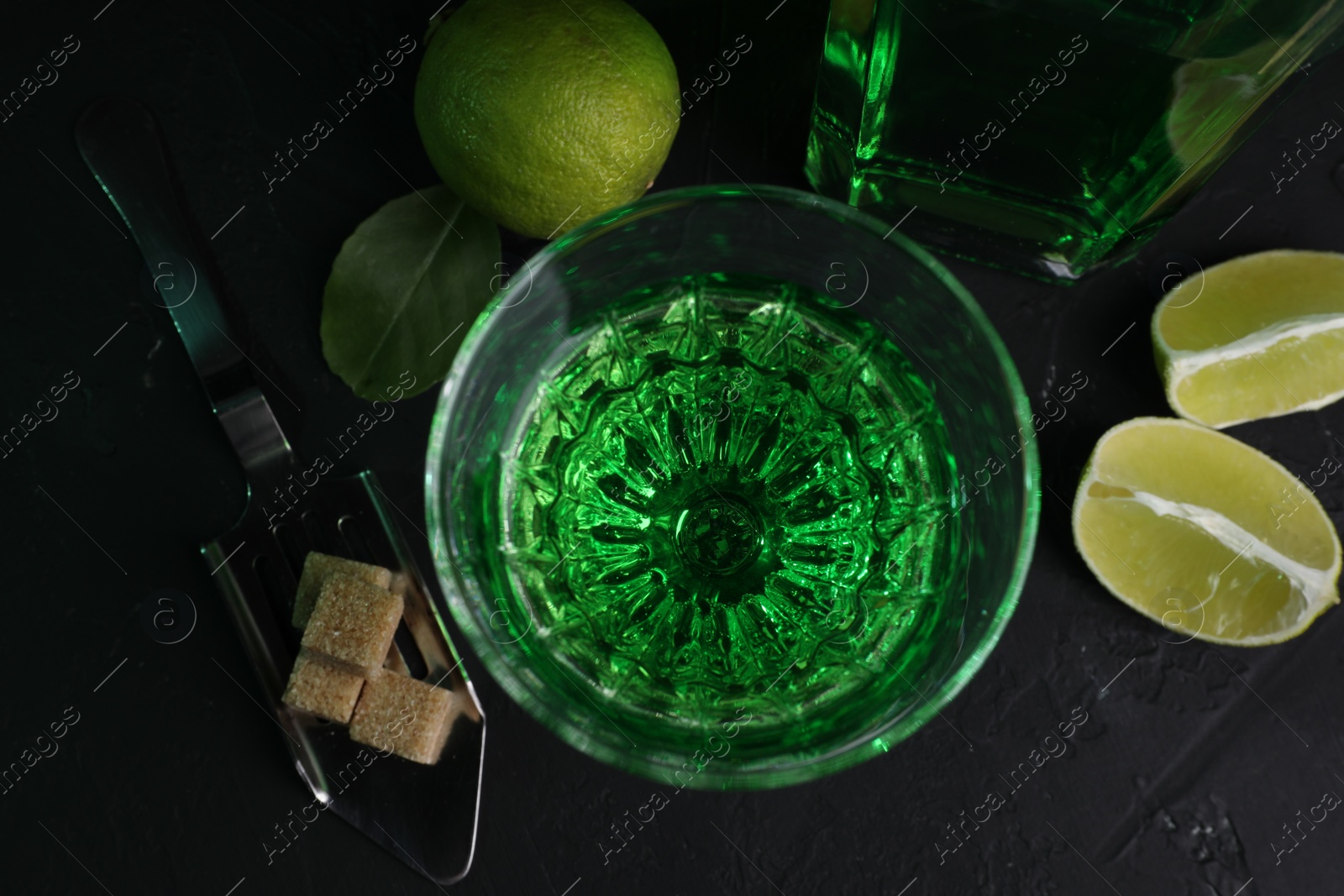 Photo of Absinthe in glass, spoon, brown sugar and lime on black table, flat lay. Alcoholic drink
