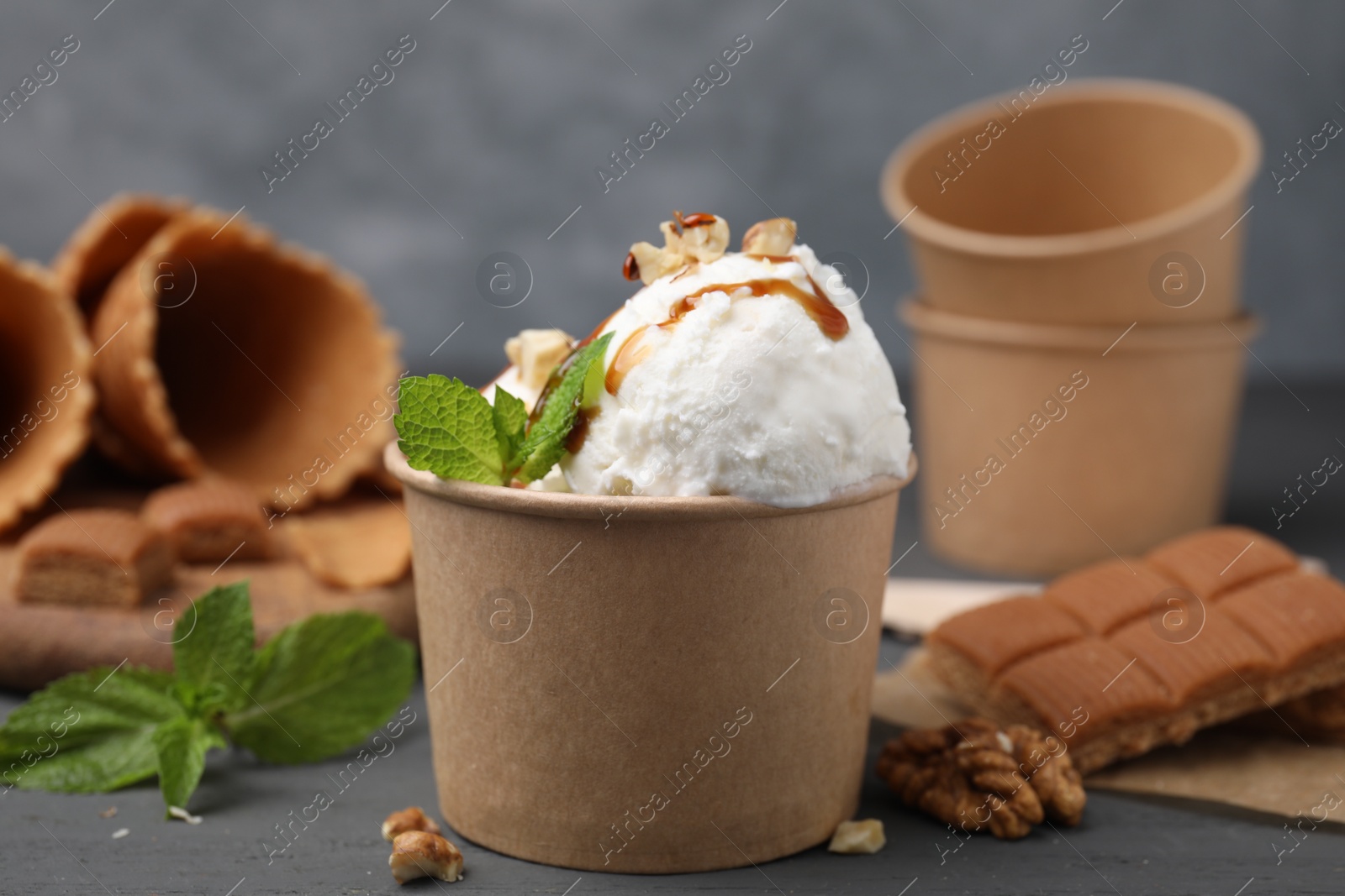 Photo of Tasty ice cream with caramel sauce, mint and nuts in paper cup on grey wooden table, closeup