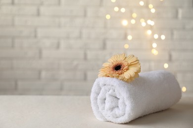 Rolled terry towel and flower on white table near brick wall indoors, closeup. Space for text