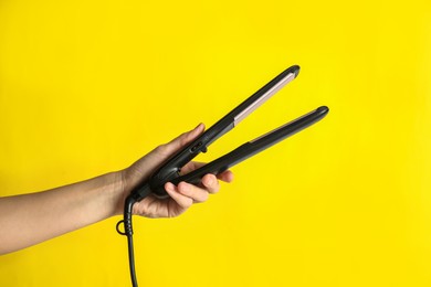 Woman holding flat hair iron on yellow background, closeup