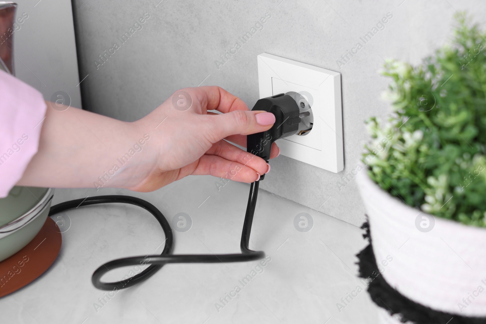 Photo of Woman plugging electric kettle into power socket at white table indoors, closeup