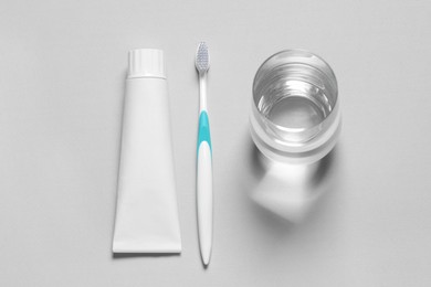 Plastic toothbrush with paste and glass of water on grey background, flat lay