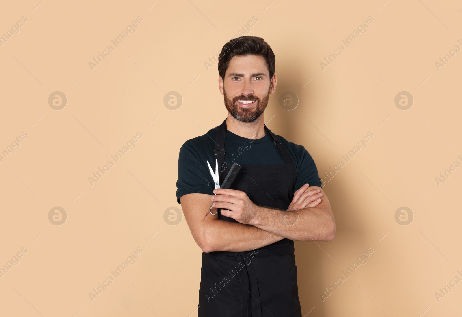 Photo of Smiling hairdresser in apron holding comb and scissors on light brown background