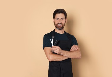 Smiling hairdresser in apron holding comb and scissors on light brown background