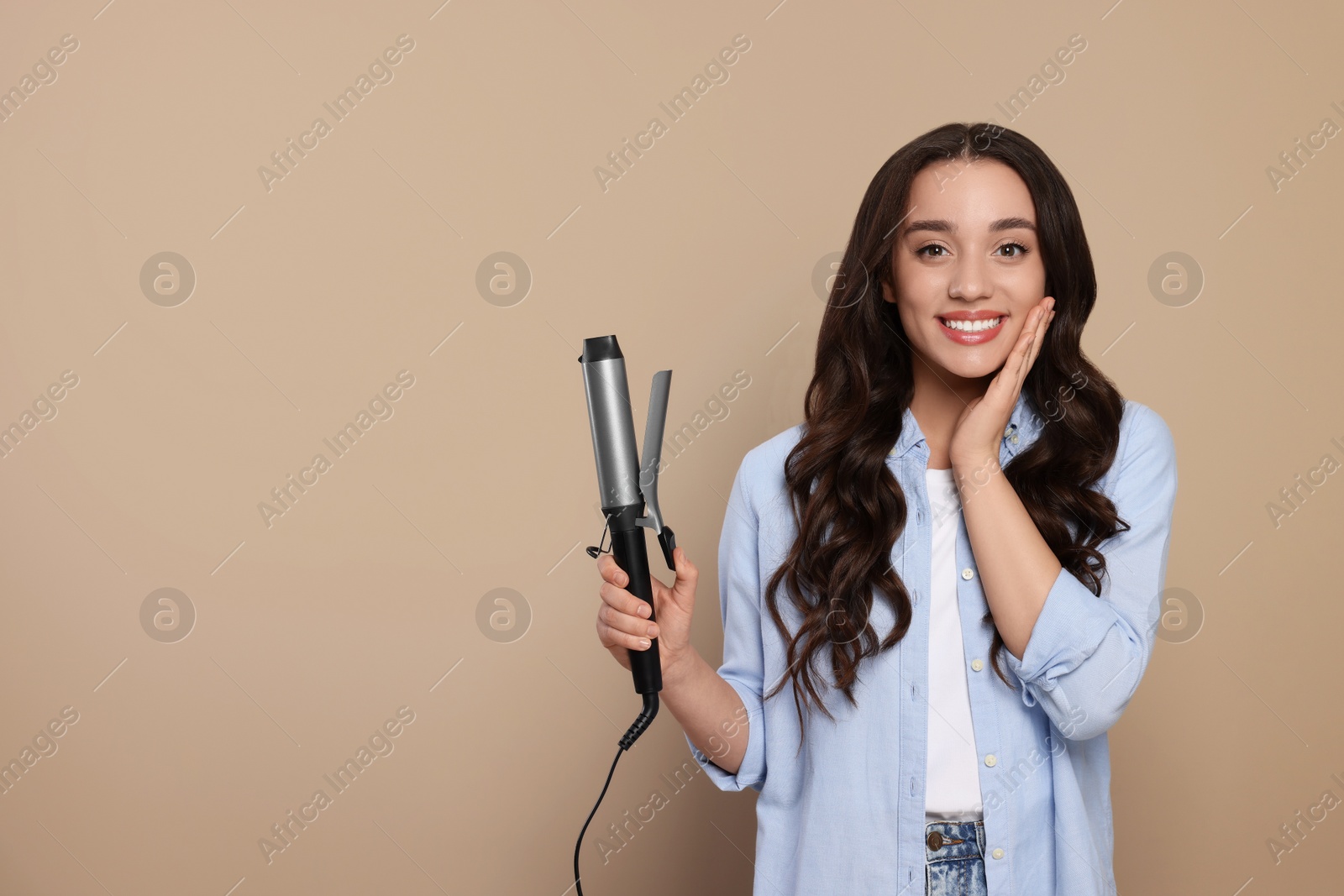 Photo of Happy woman with curling hair iron on beige background. Space for text