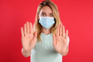 Photo of Woman in protective face mask showing stop gesture on red background, focus on hands. Prevent spreading of coronavirus