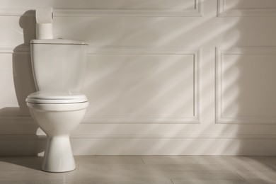 Photo of Modern toilet bowl and paper rolls near white wall in restroom, space for text. Interior design