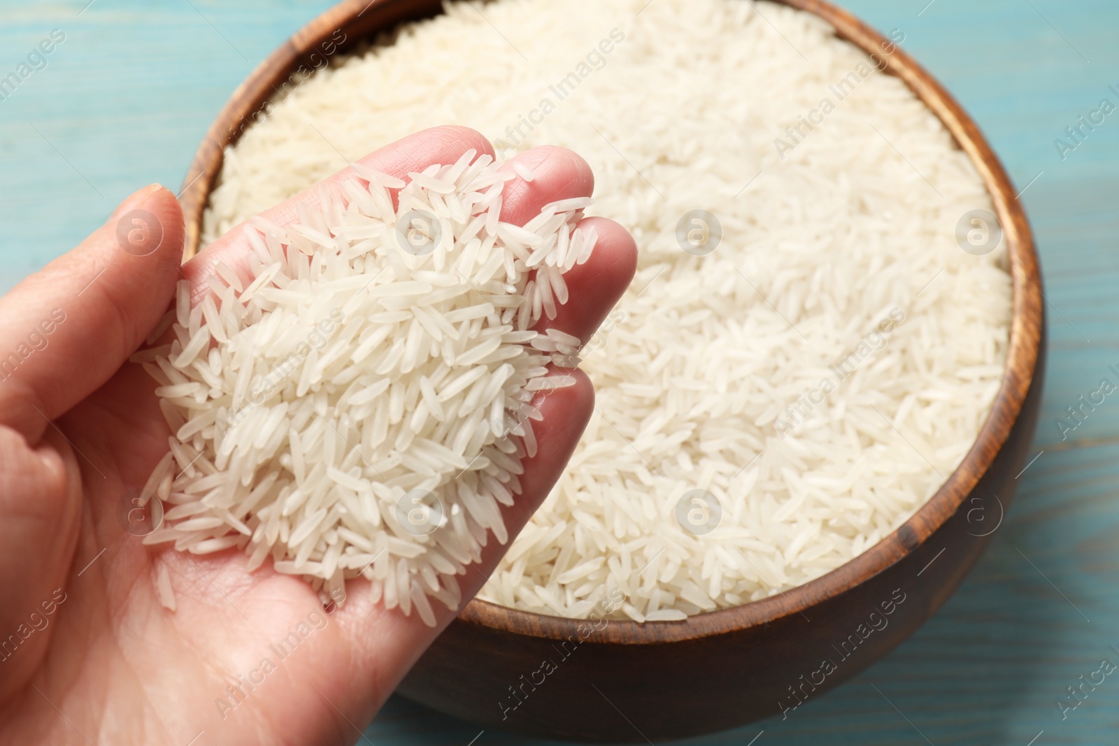 Photo of Woman holding raw basmati rice over bowl at light blue wooden table, closeup