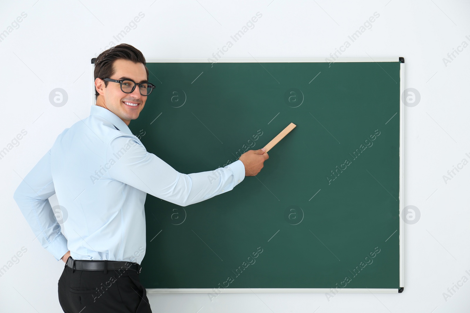 Photo of Young teacher with ruler near blank chalkboard in classroom. Space for text