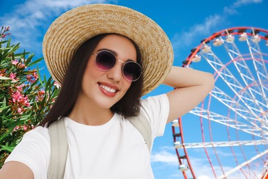 Smiling young woman in sunglasses and straw hat taking selfie near observation wheel