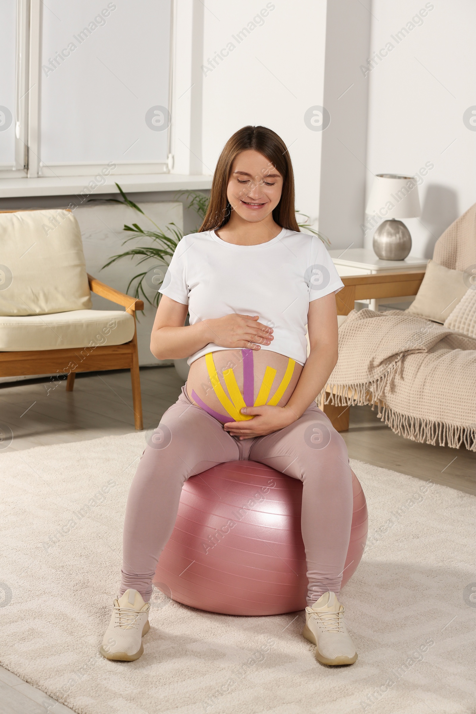 Photo of Beautiful pregnant woman with kinesio tapes doing exercises on fitball at home