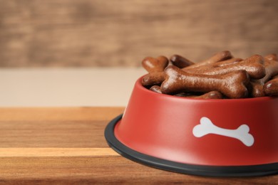 Photo of Red bowl with bone shaped dog cookies on wooden board, closeup. Space for text
