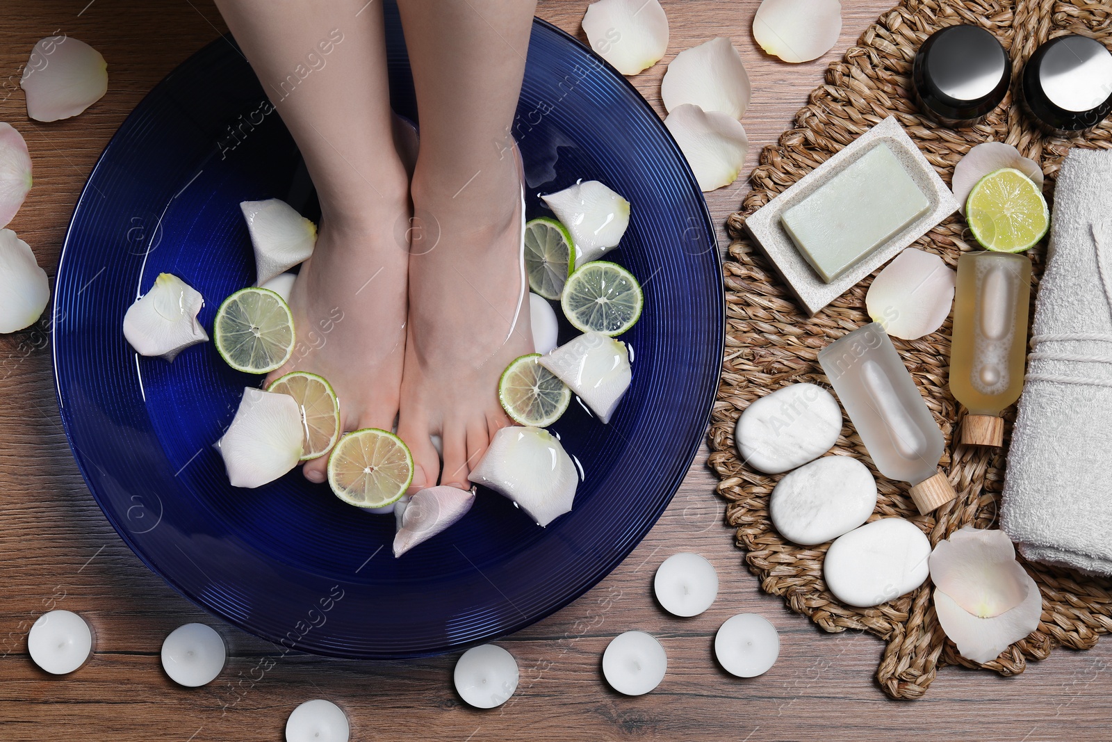 Photo of Woman soaking her feet in bowl with water, petals and lime slices on wooden floor, top view. Spa treatment