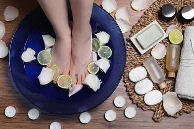 Woman soaking her feet in bowl with water, petals and lime slices on wooden floor, top view. Spa treatment