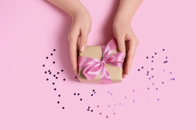 Woman holding gift box with bow and confetti on pink background, top view