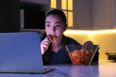 Photo of Young woman eating chips while using laptop in kitchen at night. Bad habit