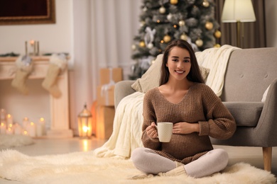 Happy pregnant woman with cup in room decorated for Christmas. Expecting baby