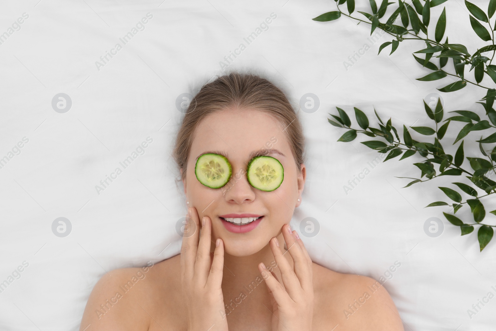 Photo of Beautiful woman with cucumber slices and leaves on white fabric, above view. Organic face mask