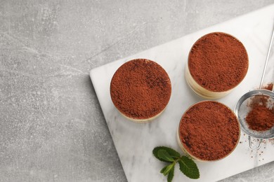 Photo of Delicious tiramisu in glasses, mint and sieve with cocoa powder on grey textured table, top view. Space for text