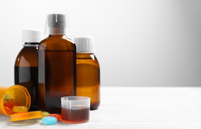 Photo of Bottles of syrup, measuring cup, lozenges and spoon on white wooden table against light grey background, space for text. Cold medicine