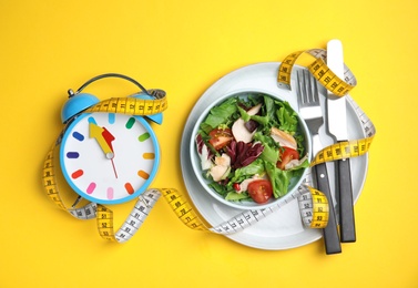 Photo of Plate of tasty salad, alarm clock and measuring tape on yellow background, flat lay. Nutrition regime