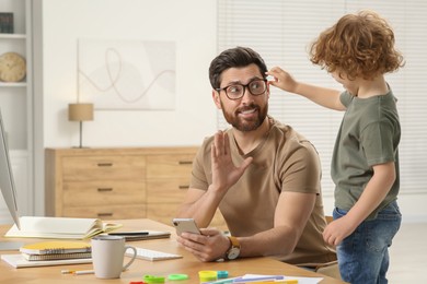 Little boy bothering his father at home. Man working remotely at desk