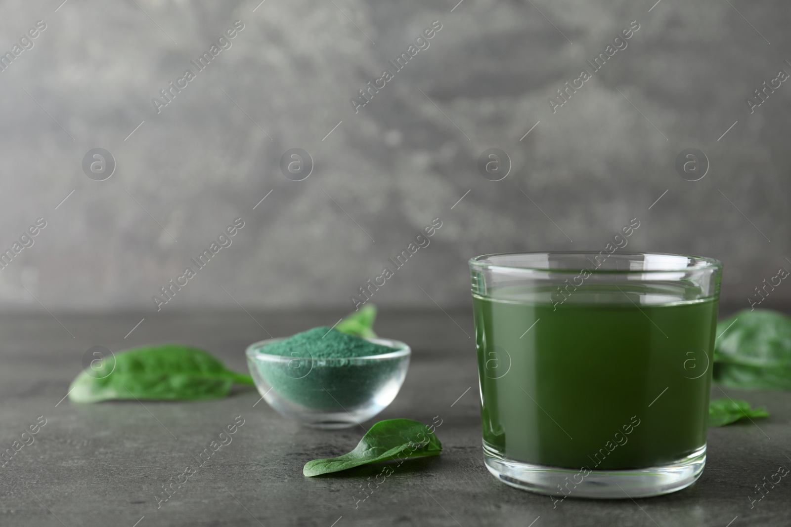 Photo of Glass of spirulina drink, spinach and powder on table against grey background. Space for text